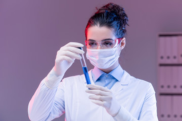 Young woman in mask, gloves and eyeglasses looking at blue substance in flask