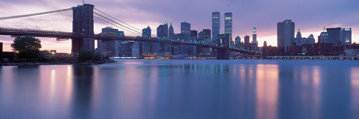 This is the Brooklyn Bridge over the East River with the Manhattan skyline at dusk. The lights of...
