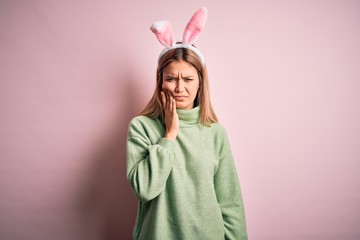 Young beautiful woman wearing easter rabbit ears standing over isolated pink background touching mouth with hand with painful expression because of toothache or dental illness on teeth. Dentist