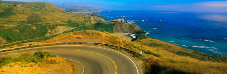 This is Route 1 also known as the Pacific Coast Highway. The road curves around a bend to the left and drops down overlooking the ocean. The rocky hillside is also seen next to the ocean. - obrazy, fototapety, plakaty