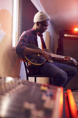Side view portrait of contemporary African-American man playing guitar sitting on high chair while...