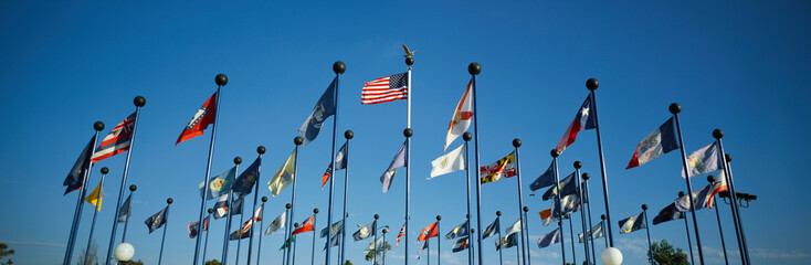 There are 50 State Flags waving in the wind on flagpoles equal distant apart against a blue sky, with the American flag in the center. These are located at Sea World. © spiritofamerica