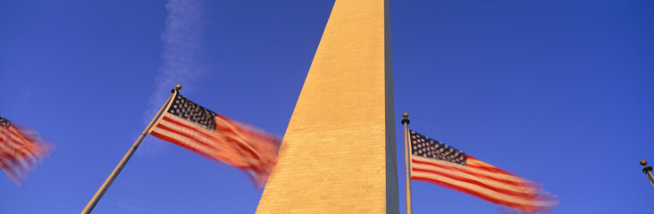 This is the center portion of the Washington Monument with American flags surrounding the Monument. It is set against a blue sky. The flags are on flag poles, waving in the wind.