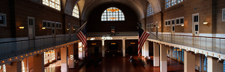 This is the interior of the Great Hall at Ellis Island which signifies immigration to the United States.  There are two American flags flying posted at the center of the walls. - obrazy, fototapety, plakaty