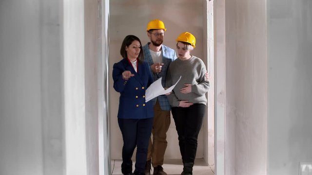 A real estate agent showing a new draft apartment to a young married couple in yellow helmets holding a layout on the paper