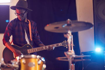 Portrait of contemporary African-American man playing guitar during rehearsal or concert with music band in recording studio, copy space