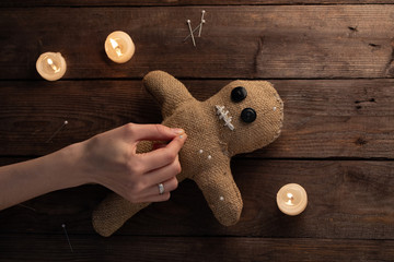 Voodoo doll on a wooden background with dramatic lighting and candles. The concept of witchcraft and black art. Burlap doll on a wooden background. Hands stick pins into a doll. Copy space.