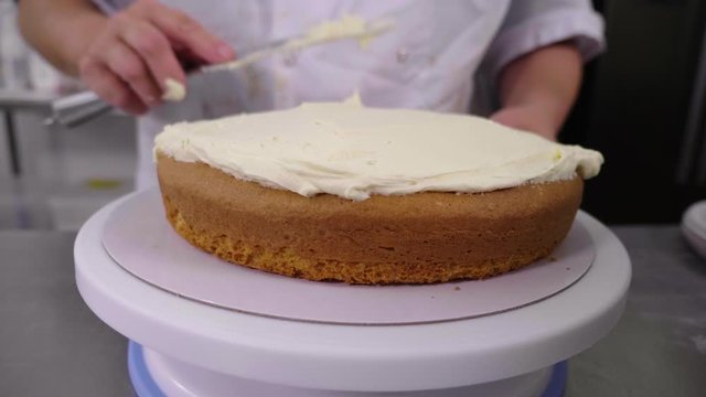 A plain sponge cake is iced with thick buttercream. A medium shot of the cake on a lazy suzy, turned by a female chef, using a palette knife to spread the mixture. A professional kitchen.
