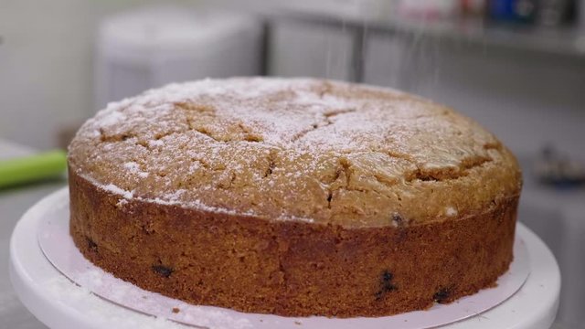 Icing sugar dusted on to a baked fruit cake in slow motion. Medium shot of cake on a lazy suzy in a well lit, professional kitchen.