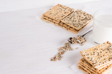 Stack of crispy wheat cakes with sesame seeds, flax and sunflower on a white wooden background.  glass of milk and spoon with sunflower seeds in the background. vegetarian food, eco food concepts