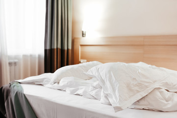 Fototapeta na wymiar crumpled white factory bedding on the bed with a blanket and pillows, next to the bedside table and dark heavy curtains on the floor, a standard elegant look of a hotel room