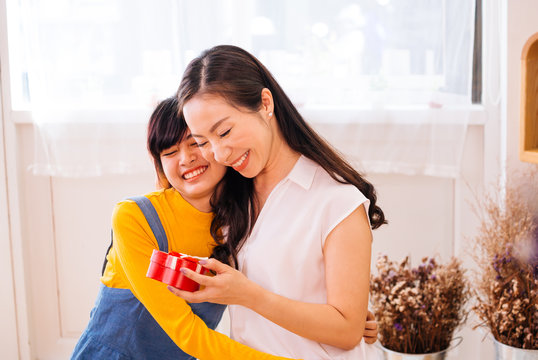 Smiling happy Asian teenage daughter and Asian middle-aged mother hugging together in indoor living room at home. Mum is holding a present gift.