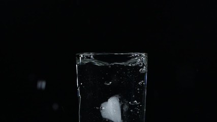 Slow motion close-up of ice cube falling down in a glass of water on black background with splash, bubbles and drops - Powered by Adobe
