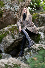 Naklejka na ściany i meble beautiful European girl in a brown plaid wool coat, scarf, leather tight pants, boots sits on huge stones covered with moss in the park