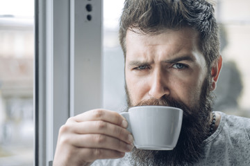 Grumpy looking bearded man drinking morning cup of coffee near window. Hipster man having cup of americano. Morning routine with coffee and start of new day. Coffee addicted.
