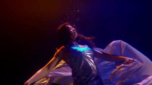 a brunette with long hair and a silver dress and leggings floats underwater in the dark. she leans back and floats up. blue and red backlight