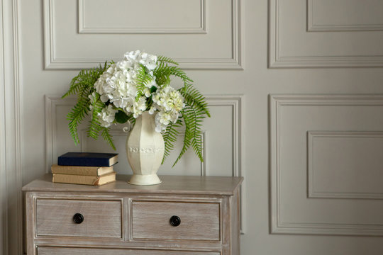 A white vase with white flowers stands on the dresser against the wall.