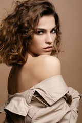 beauty fashion model with clean skin and curly hair in biege cloak stretch with scarf on biege background, bare shoulders