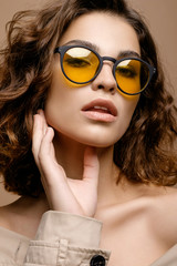 closeup portrait of beauty fashion model with clean skin and curly hair in biege cloak stretch on biege background, model in fashion glasses, bare shoulders
