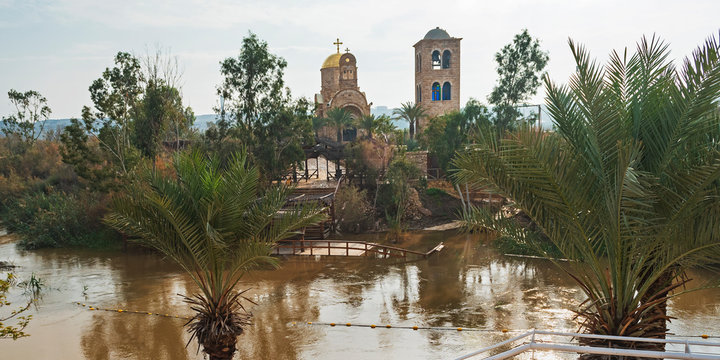 site of the baptism of jesus on the jordan river showing ancient churches in the background with palm trees and border markers in the foreground