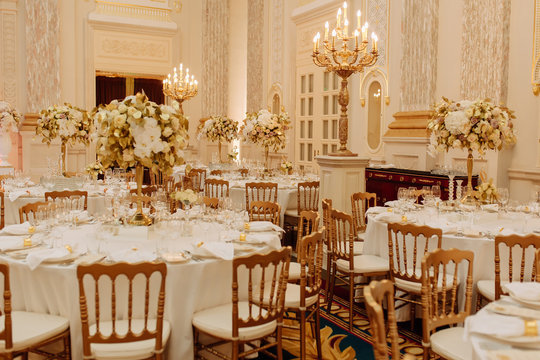 Fancy Interior Of Restaurant Celebrating Room. Dining Table With Wooden Chairs Decorated Pastel Beautiful Flower Bouquet And Dishware On White Cloth, Magnificent Golden Chandeliers