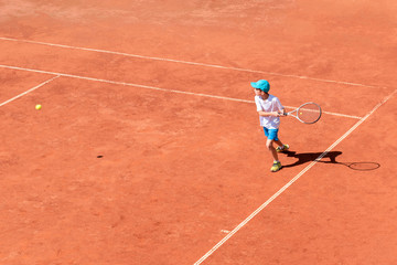 Boy tennis player plays on clay tennis court. The child is trying to hit the backhand, focused on...