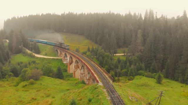 Train on the railway bridge in the Carpathian mountains. Picturesque alpine landscape with train on old arch bridge on green forest background.