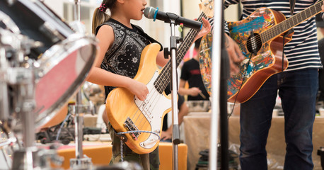 Fototapeta na wymiar Beautiful, talented young Asian girl sings and plays natural wood color electric bass guitar on contest stage with passion, joy and confidence. Indy family perform, father on guitar, brother on drums.