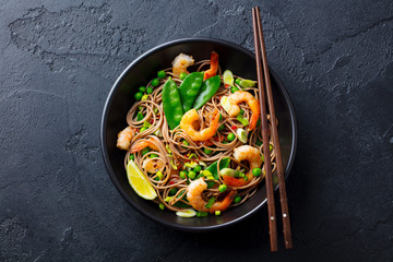 Stir fry noodles with vegetables and shrimps in black bowl. Slate background. Top view.