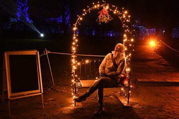 Fototapeta na wymiar Girl on a bench. Night scene with young woman seated on bench under mistletoe in night lights. Shiny Christmas light garlands on arch in darkness. Bright orange color floodlight. Christmas tradition
