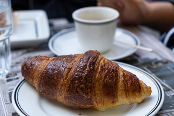 A well-done croissant on white dish in close up view. Blur cup on a saucer background.