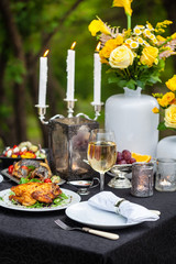 Baked full chicken with vegetables in a plate on a table next to glass of wine and candles. Outdoor, green park