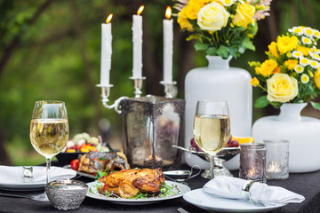 Baked full chicken with vegetables in a plate on a table next to glass of wine and candles. Outdoor, green park
