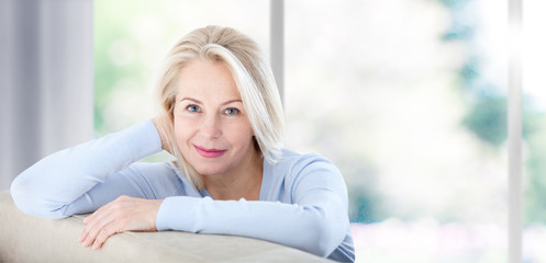 Active beautiful middle-aged woman smiling friendly and looking in camera near window at home....