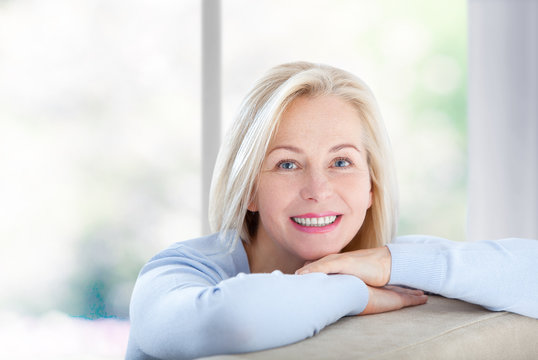 Active beautiful middle-aged woman smiling friendly and looking in camera near window at home. Woman's face closeup. Selective focus.