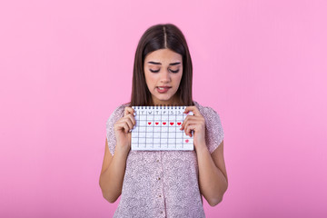 Portrait of embarrassed cute girl biting her lip and holding menstrual calendar with drawn hearts for period and looking at camera isolated over pink background