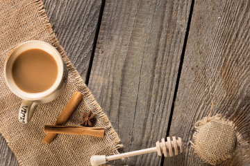 Cup of coffee on jute material with cinnamon stick, anise star, honey and dipper on wood rustic plank. Vintage background with copy space.