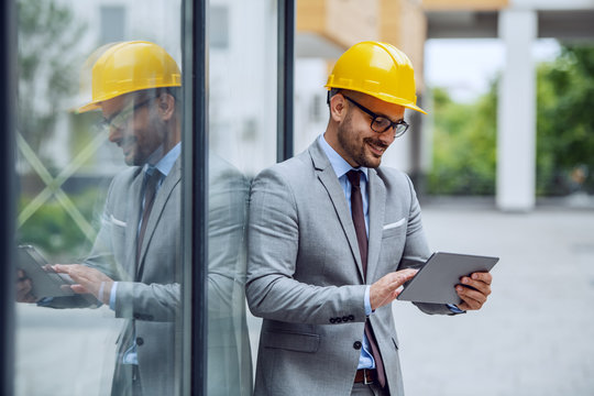 Handsome caucasian smiling elegant architect in suit, with eyeglasses and helmet on head leaning on window at construction site and using tablet.