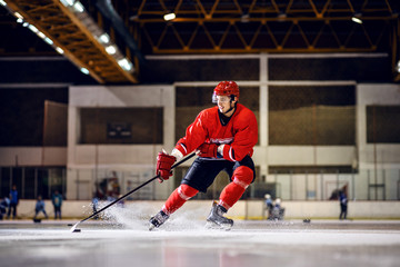 Strong Caucasian hockey player in red uniform with helmet skating to goal with stick and puck.