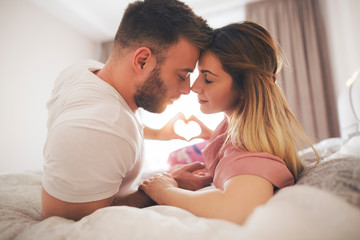 Beautiful couple in bed showing heart symbol with their hands.