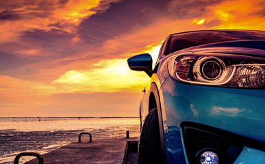 Blue compact SUV car with sport and modern design parked on concrete road by the sea beach at sunset. Front view of luxury car. Closeup SUV fog lamp and headlights with beautiful sunset sky in summer.