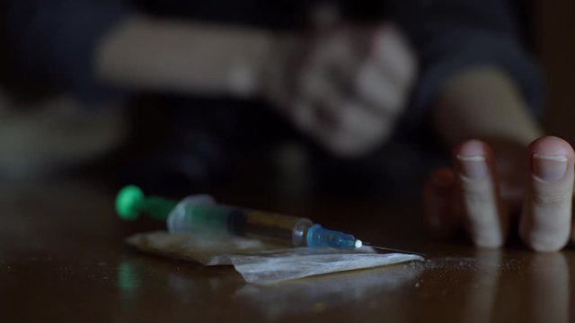 A female drug addict reaches out to a syringe and a bag of powder. Does himself an injection or injection into the vein of the arm.