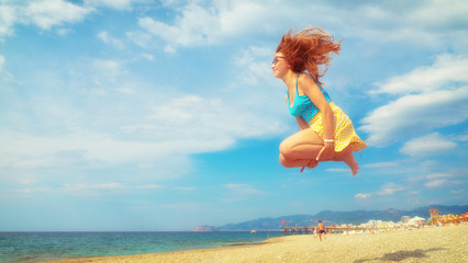 Naklejka premium Excited young woman having fun on beach in summer. Side view of carefree youthful female tourist in sunglasses enjoying freedom and jumping on sandy seashore