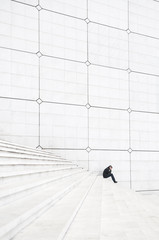 Distant figure of a man huddling with his head in hands looking very small on a large white staircase