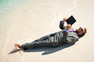Relaxed businessman lying in a wet suit using his tablet computer on the shore of a tropical beach