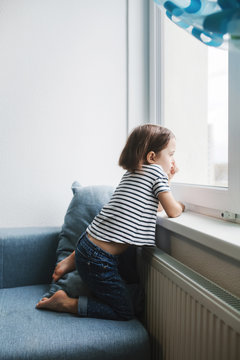 Little Bored Girl Sitting On The Couch At Home And Looking At The Window