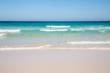 Naklejka premium Cloudless sky over a colorful sea with diferent colors aquamarine turquoise dakr blue azure with waves prducing white foam crushing against a yellow beach summer sumertime sunny calm water tranquil