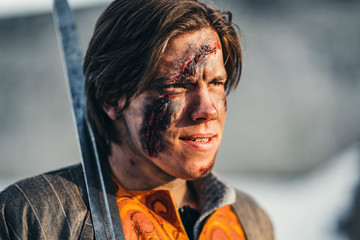 Portrait of a young medieval warrior in armor with sword on his shoulder. In the process of battle, with wounds on the face