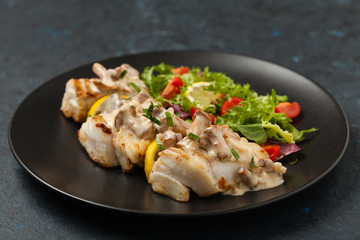 Fried cod served with mushroom sauce and salads. Portion on a black plate. Dark background.