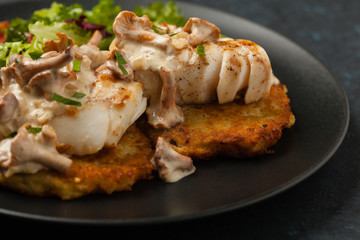 Fried cod, served on potato pancakes with mushrooms sauce and salads. Portion on a black plate. Dark background.